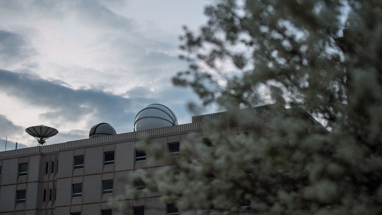 The Van Allen Observatory dome is pictured atop Van Allen Hall along with other roof structures as viewed from the southeast. A tree with spring growth partially obscures the view.
